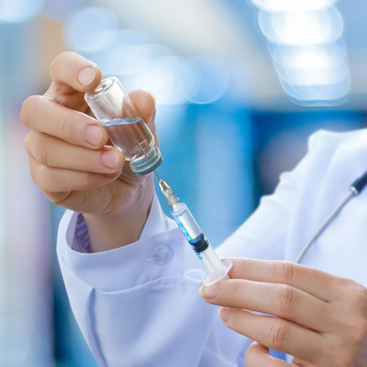 Close-up of a healthcare professional drawing liquid from a small glass vial into a syringe, with a blurred clinical background.