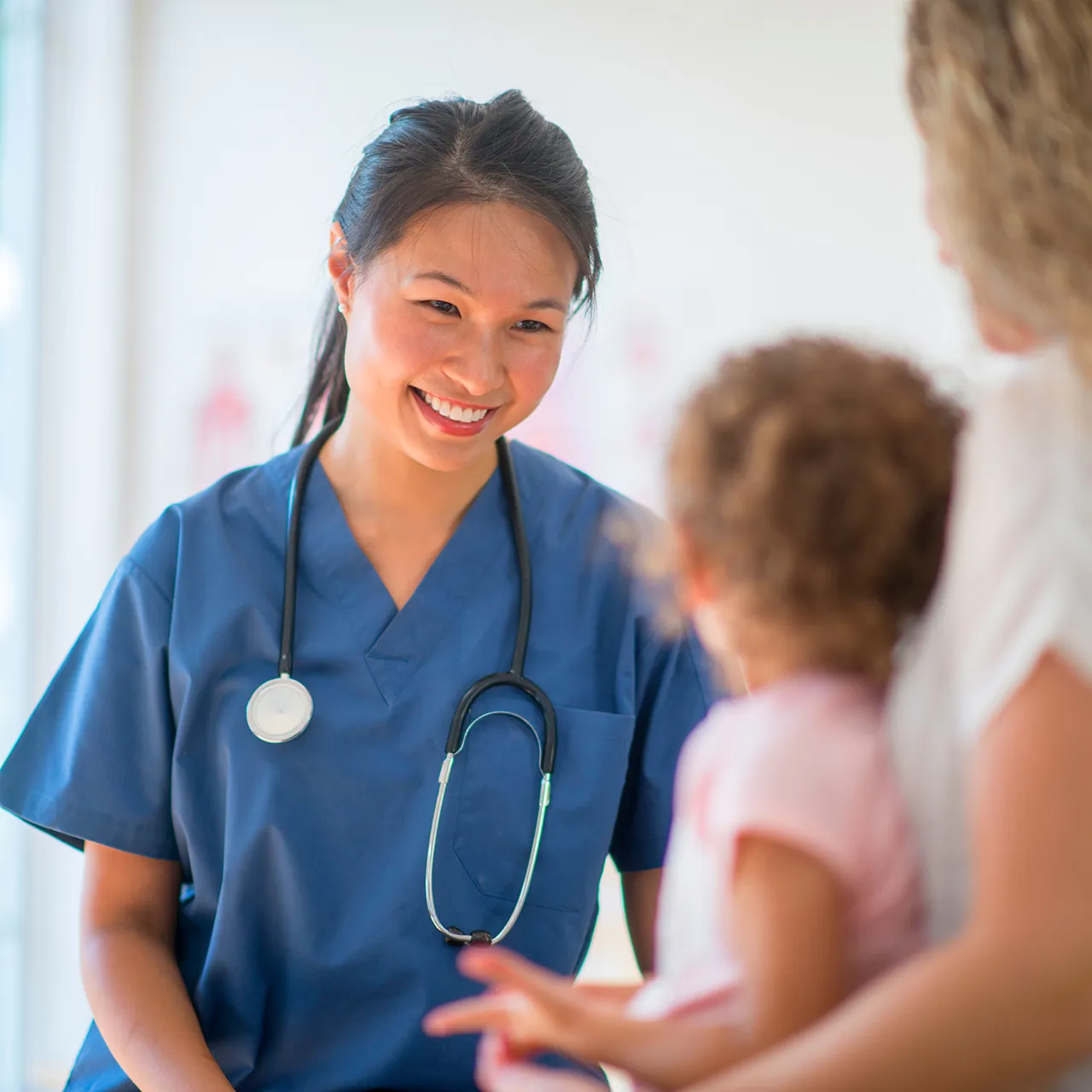 Healthcare professional in blue scrubs with a stethoscope smiling while speaking to a young child and parent in a clinical setting.