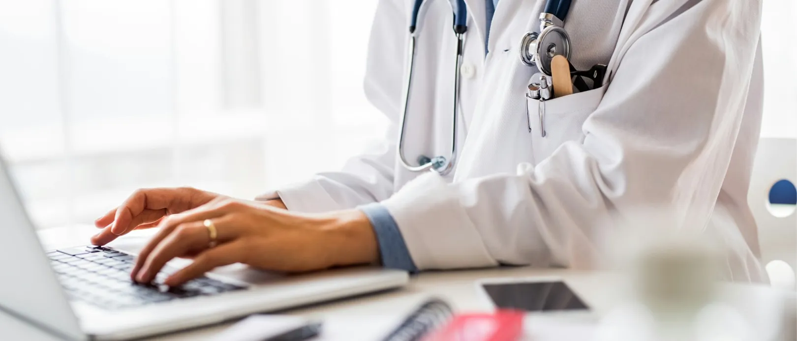 A woman in a white coat is sitting at a desk, working on her laptop.