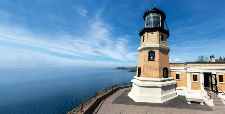 Image of Split Rock Lighthouse, up close. It appears to be up on the top of a hill, overlooking water and the sky, and there's a couple story tall lighthouse that is 20 feet away.