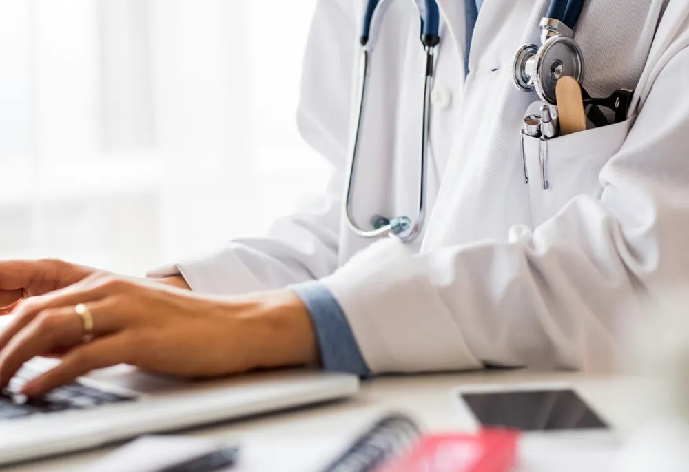 A woman in a white coat is sitting at a desk, working on her laptop.