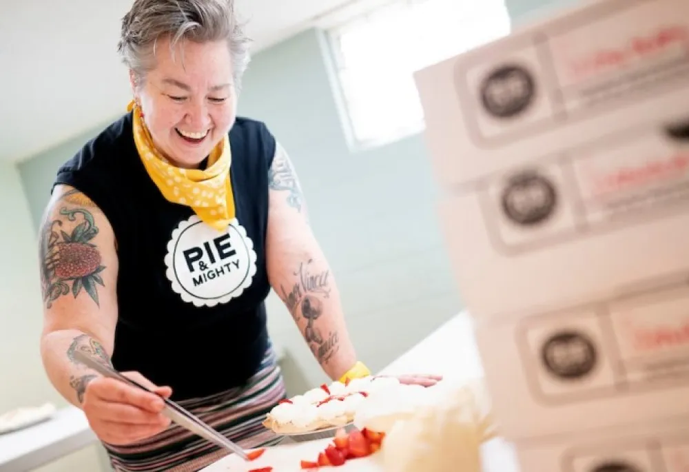 Image of a person (Rachel, one of the owners of Pie & Mighty) using a tweezers to place strawberries as garnish on a pie. There's also a stack of pie boxes in the foreground. 
