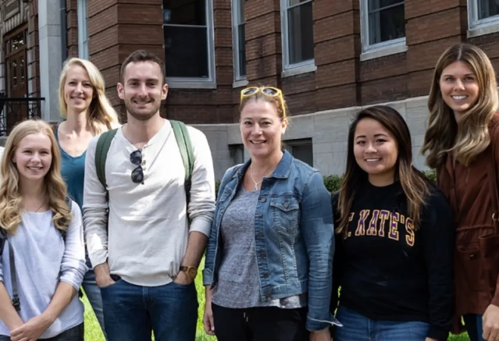 A group of college students, mostly women, standing in front of a brick college building, smiling. 