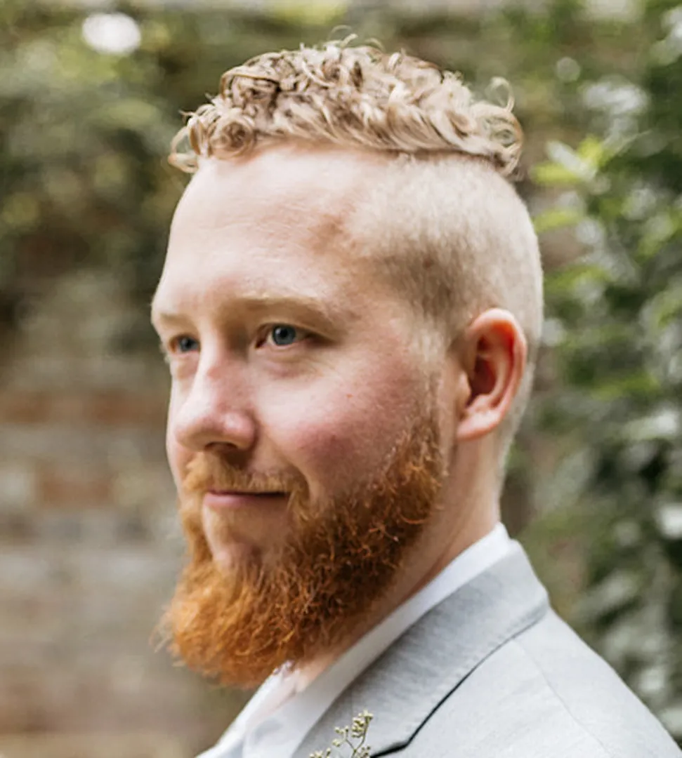 John Doyle - Side view of a man with short hair on the sides and a dapper beard. Background of blurred trees.