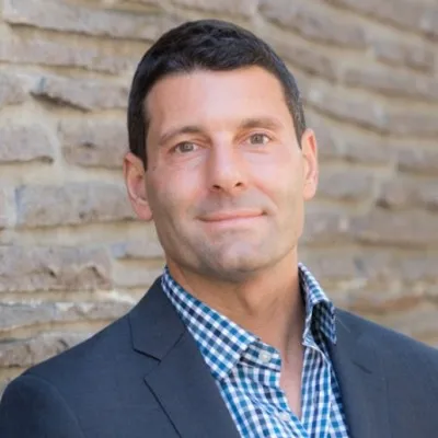 Scott Delea - Clean shaven man in a blue sport jacket and blue checked shirt. He is in front of an exposed brick wall.