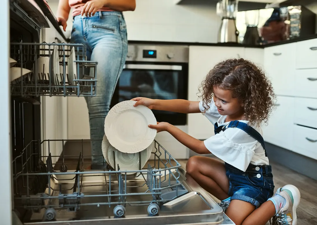 Child packs the dishwasher while her parent stands in the background