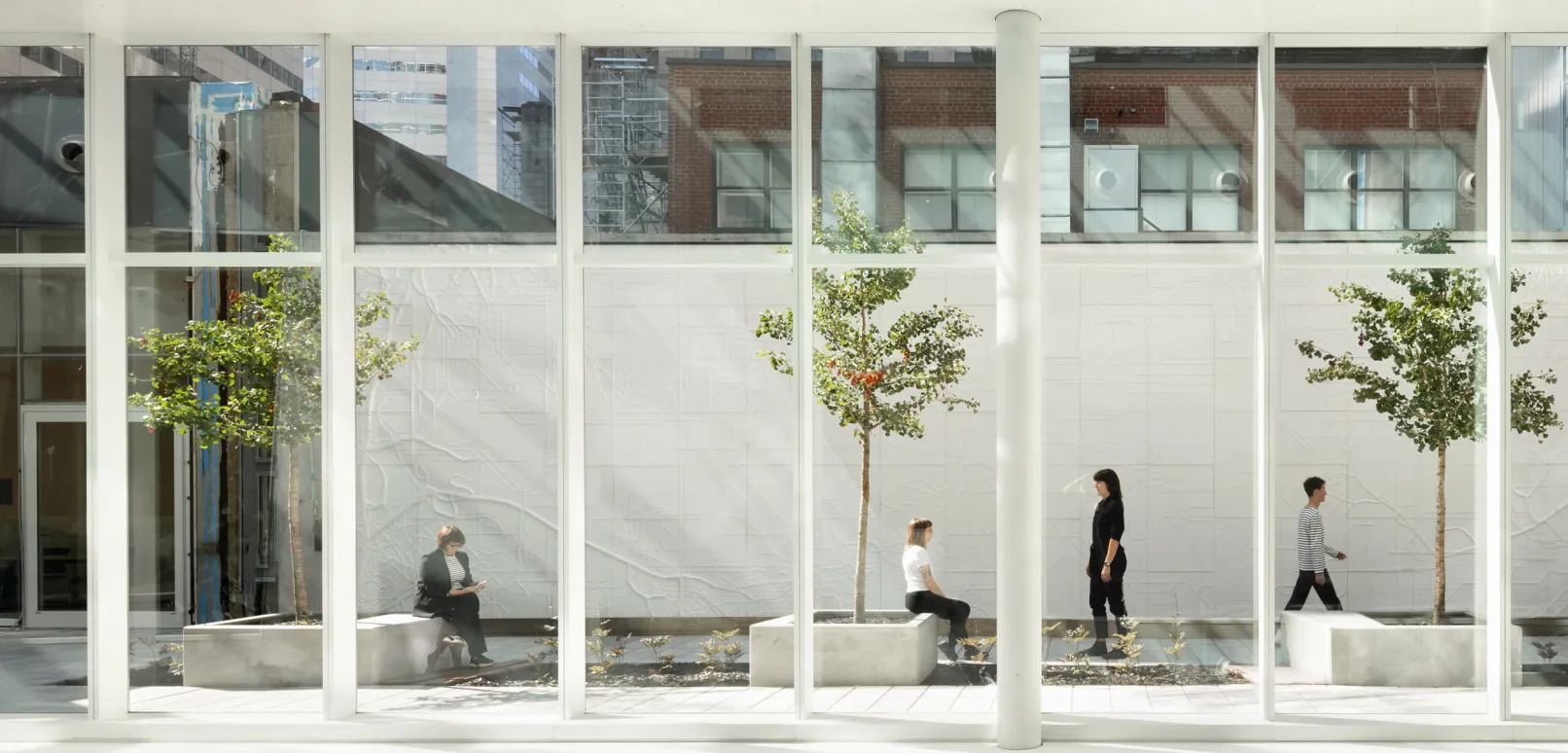 Bright modern atrium with floor-to-ceiling windows, indoor trees, and people sitting, standing, and walking along a white tiled courtyard.