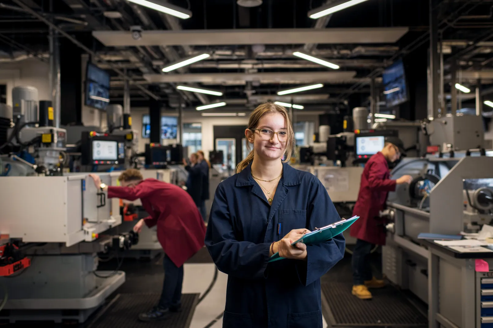 Engineering student holding a folder in a university workshop, with other students and equipment behind her