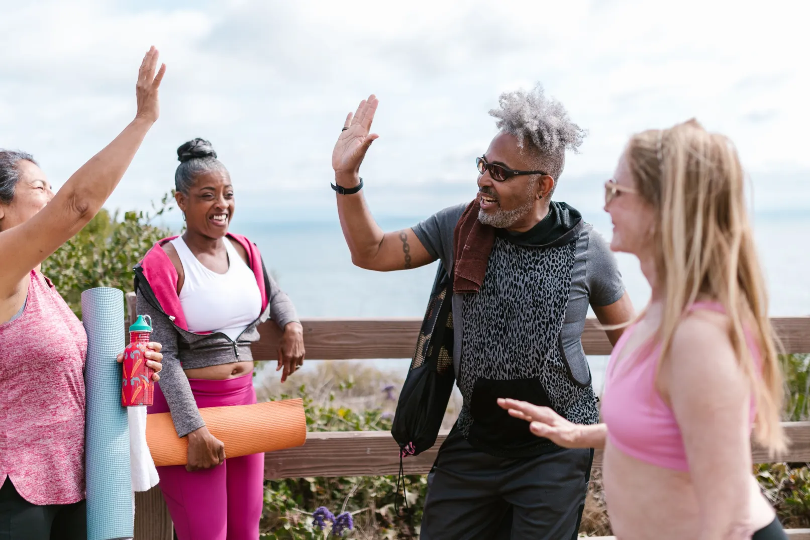 A group of adults with exercise mats smiling and raising their hands outdoors