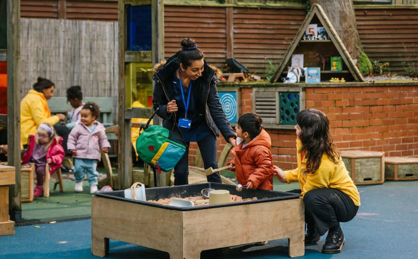 A parent and child with a childcare provider beside a sandpit in an outdoor nursery play area