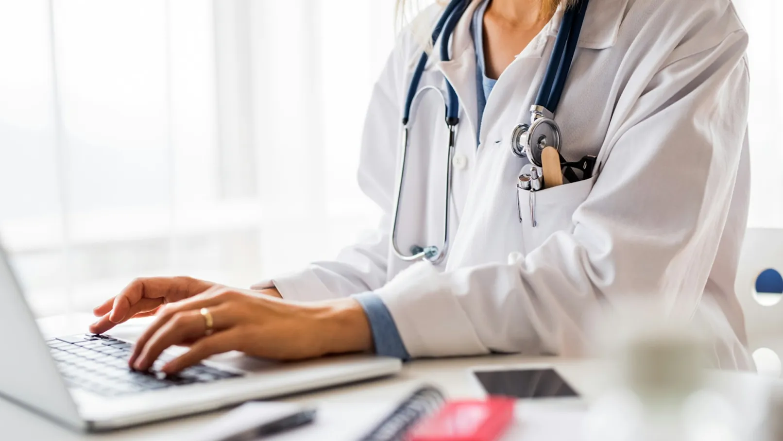 A woman in a white coat is sitting at a desk, working on her laptop.