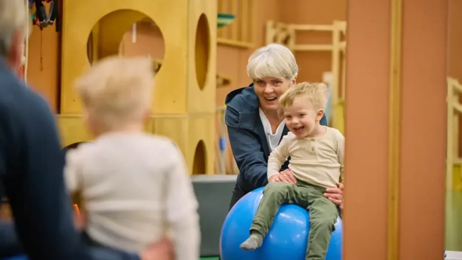 An older lady holds a smiling child who sits on top of a large blue exercise ball. 