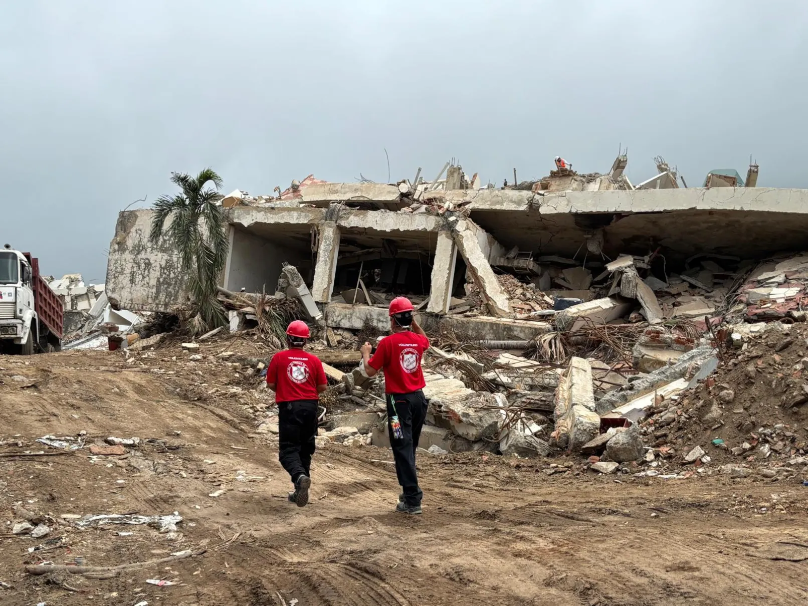 Two Salvation Army volunteers wearing red shirts and helmets walk towards a collapsed building