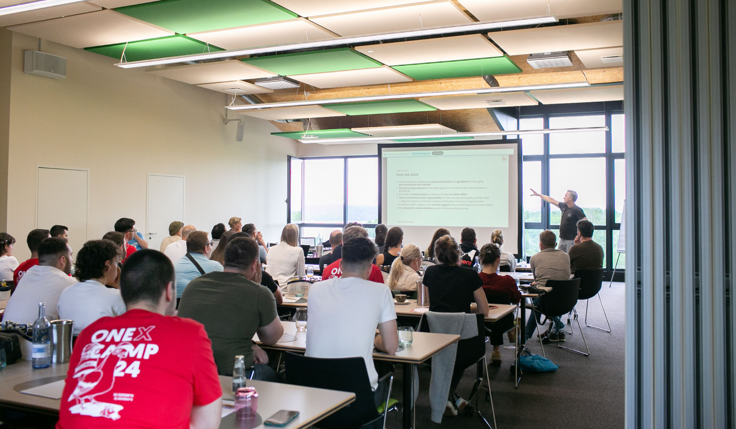 A room full of  people watch a person give a presentation in front of a big screen.
